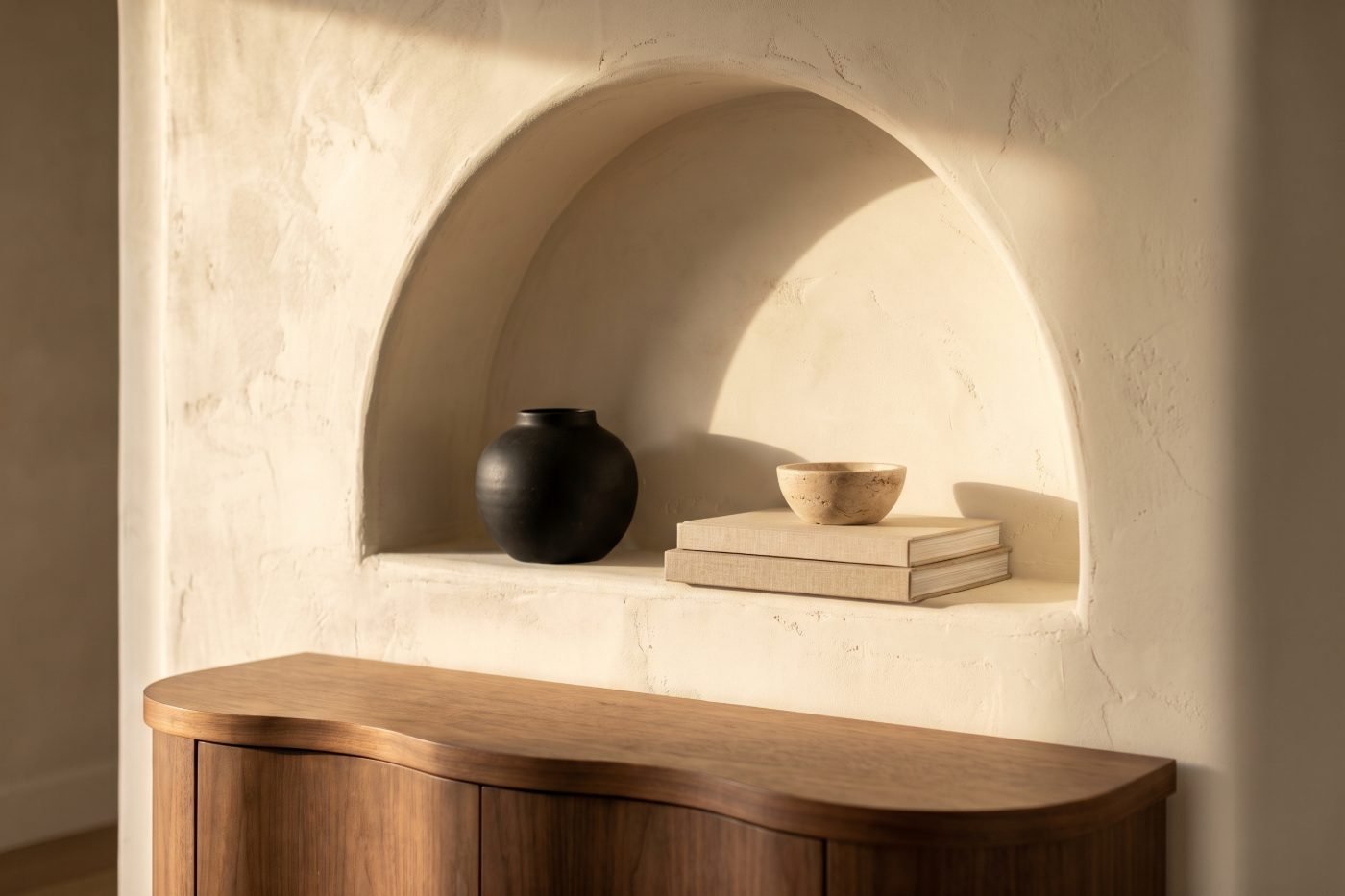 Arched plaster wall niche holding a black ceramic vessel and travertine bowl above a curved walnut console.