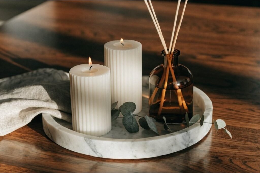 Two lit ribbed ceramic candles and an amber glass reed diffuser on a marble tray with eucalyptus