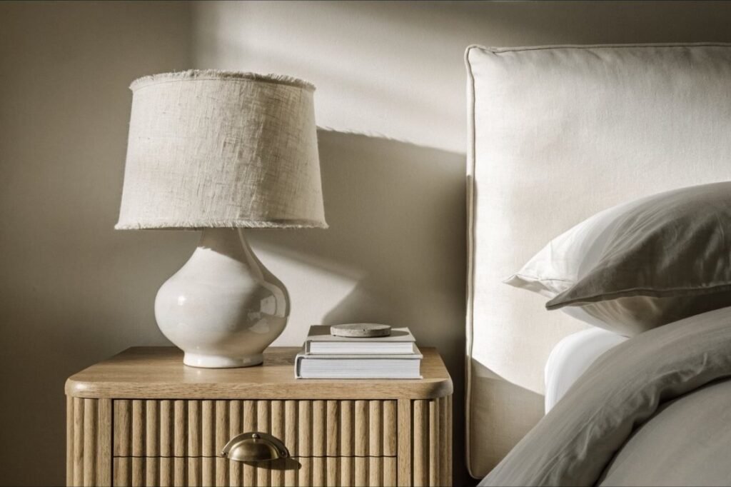 A fluted oak nightstand with a brass pull, ceramic lamp and books beside a cream upholstered headboard