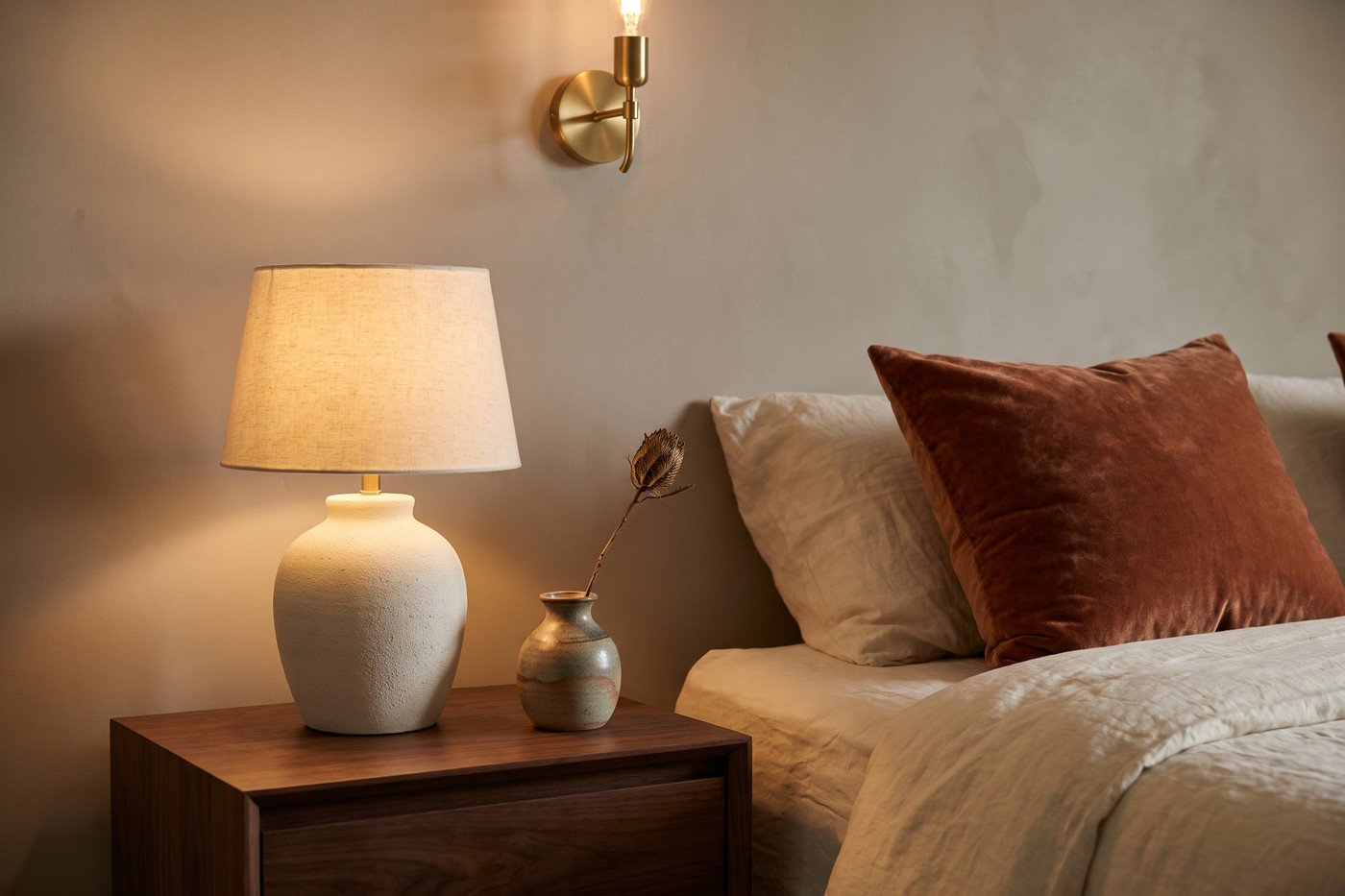 Bedside vignette with a walnut nightstand, glowing ceramic lamp, clay velvet pillow and brass sconce on a putty wall.
