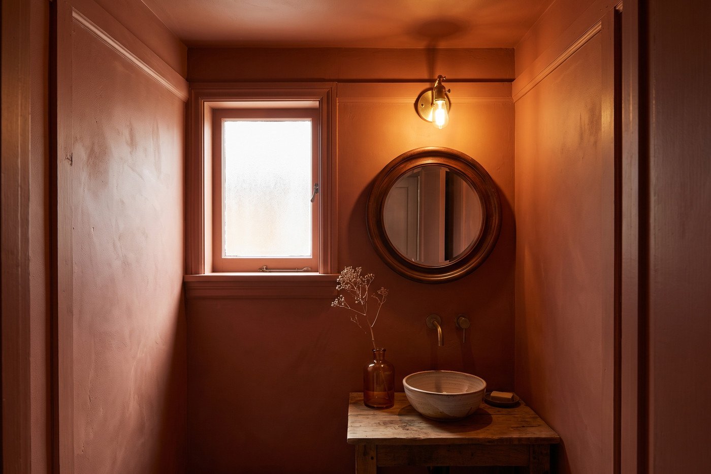 Small powder room colour-drenched in warm clay terracotta with a round wood mirror, brass sconce and amber glass vase.