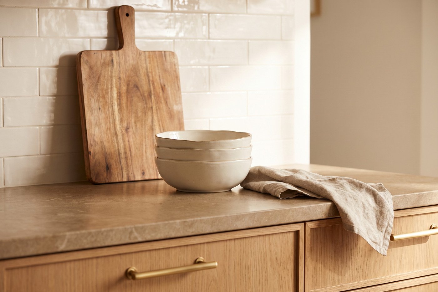 Kitchen counter vignette with stacked cream stoneware, an acacia serving board and brass cabinet pulls on oak drawers.