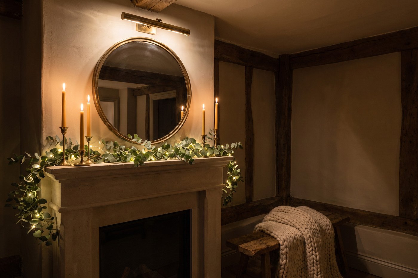 Fireplace at night with lit tapers on the mantel, warm string lights woven through a greenery garland, a rechargeable picture light above and a chunky knit throw on the hearth.