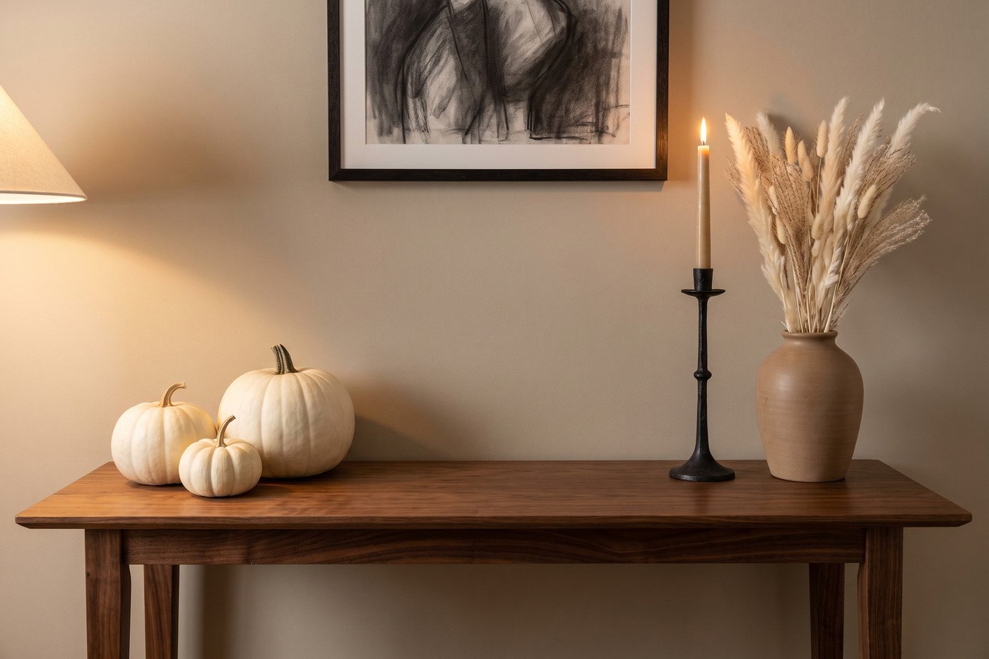 Console table with three white pumpkins at different sizes, a black candlestick, a tall ceramic vase of dried white stems and a dark framed artwork above.