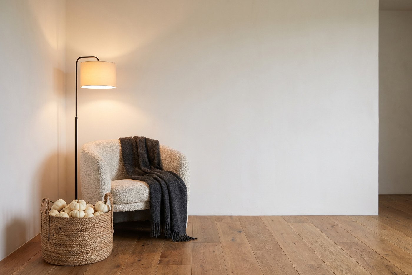 Reading corner with a cream bouclé chair, a charcoal wool throw, a black floor lamp lit warm and a woven basket of white pumpkins on the floor.