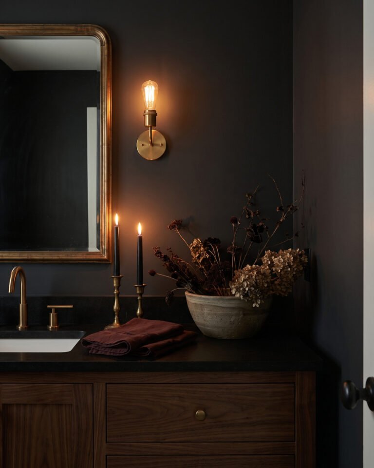 Moody guest bathroom styled for Halloween with charcoal walls, an antique brass mirror, black taper candles in brass holders on the vanity, dark burgundy hand towels and a low bowl of dried black-stemmed flowers.