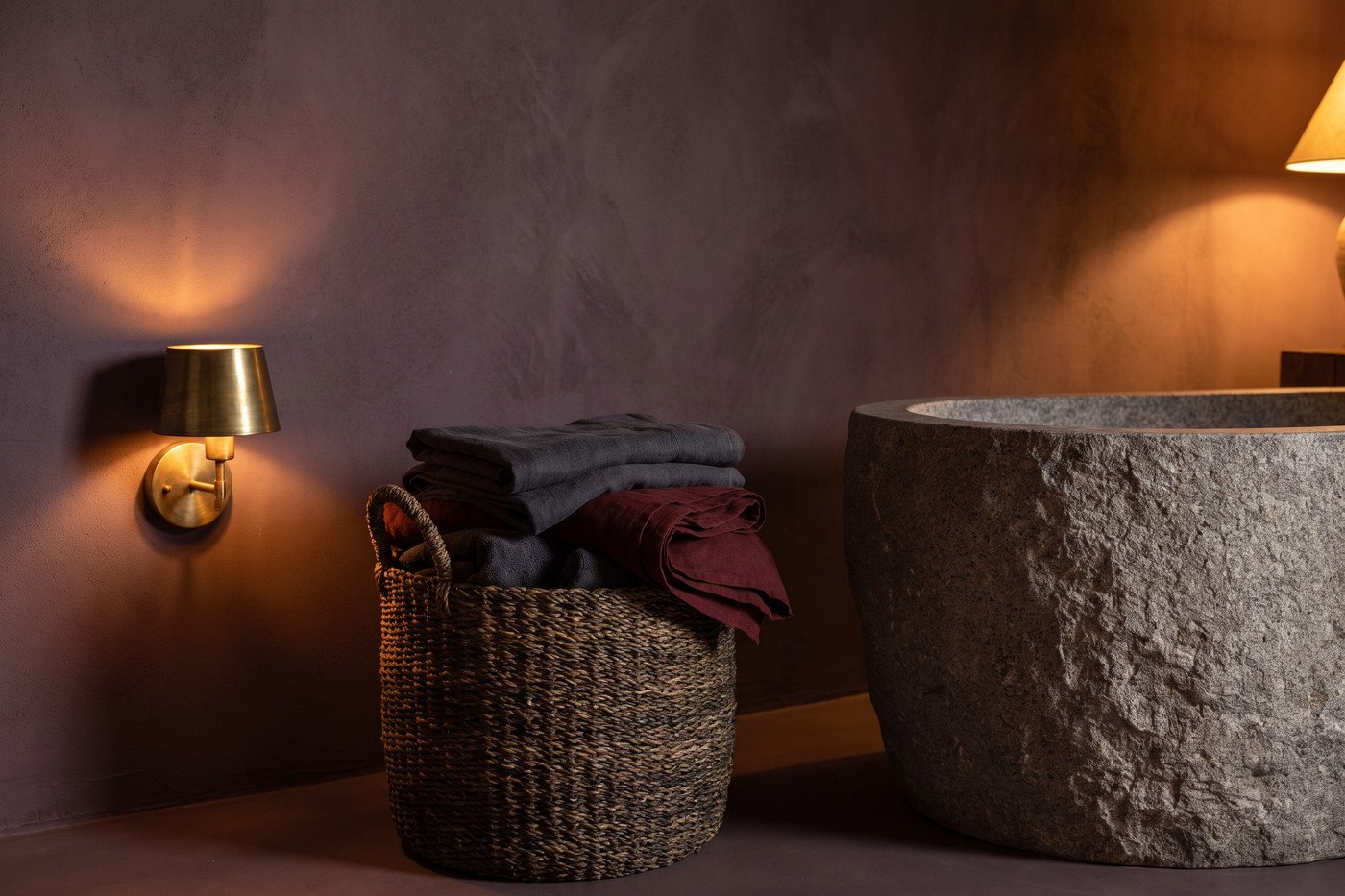 Stacked charcoal and oxblood towels in a woven basket beside a freestanding tub, with a brass sconce lit low on a deep plum wall.