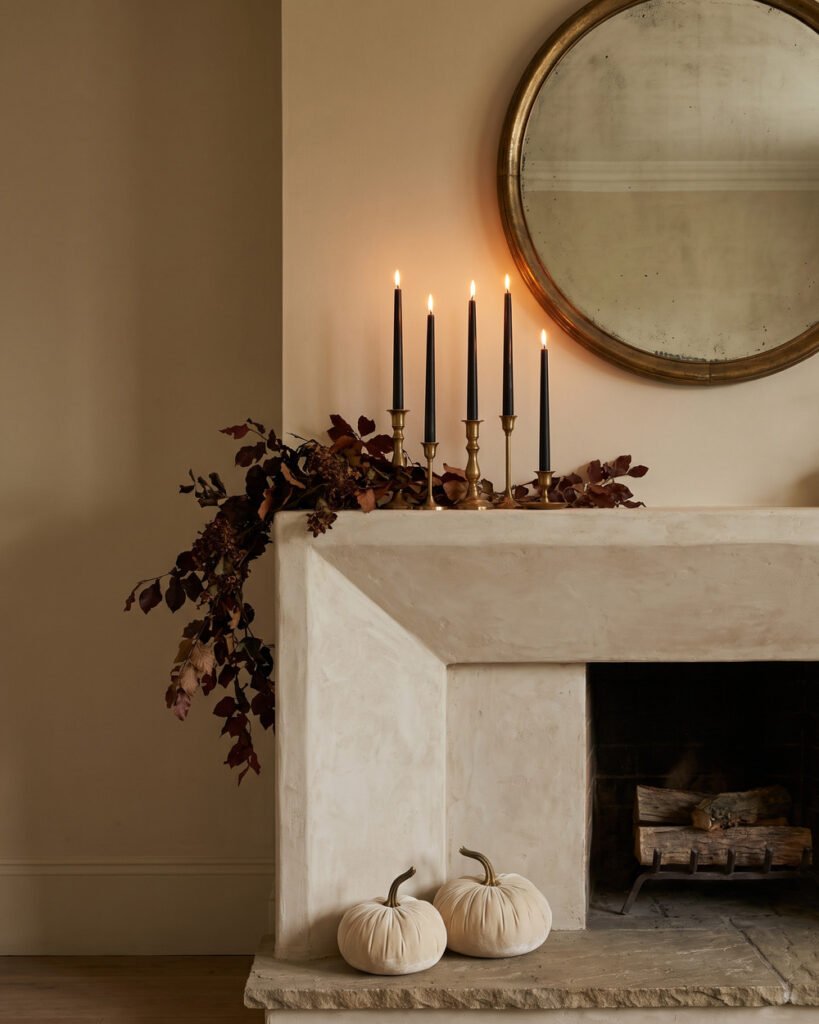 Limewashed fireplace mantel styled for Halloween with black taper candles at five heights in brass holders, a dark dried garland trailing off one end, cream velvet pumpkins and an antique mirror above.