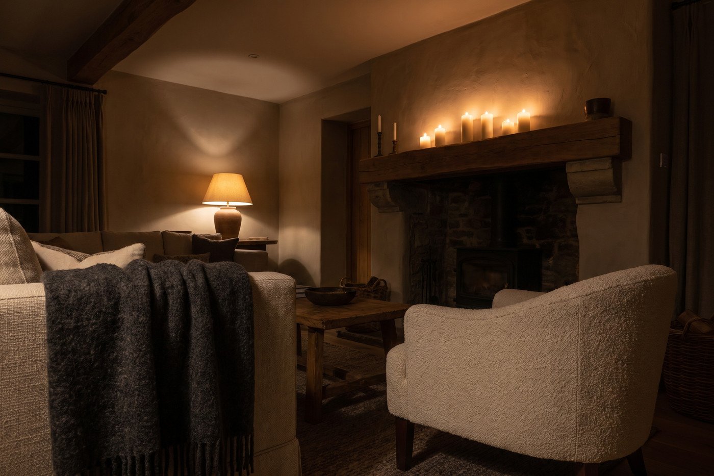 The fireplace seen from the room at night, mantel candles lit, a wool throw over a bouclé chair and warm lamplight in the corner, the ceiling light off.