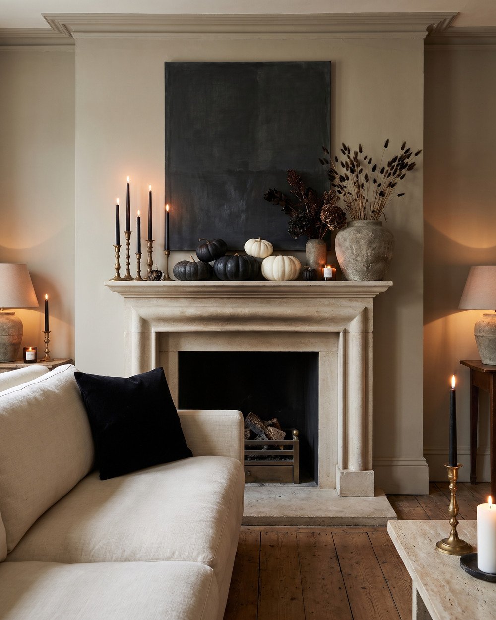 Elegant Halloween living room with a limestone mantel styled in black taper candles, white and black pumpkins, dried dark botanicals in a stone vase and a large moody abstract artwork, warm candlelight.