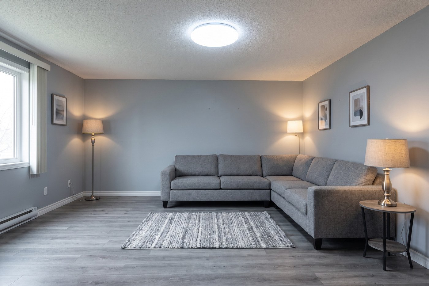 Cool grey living room with a grey sectional, grey walls, a small rug and a bright cool-white ceiling light, looking flat and dated.