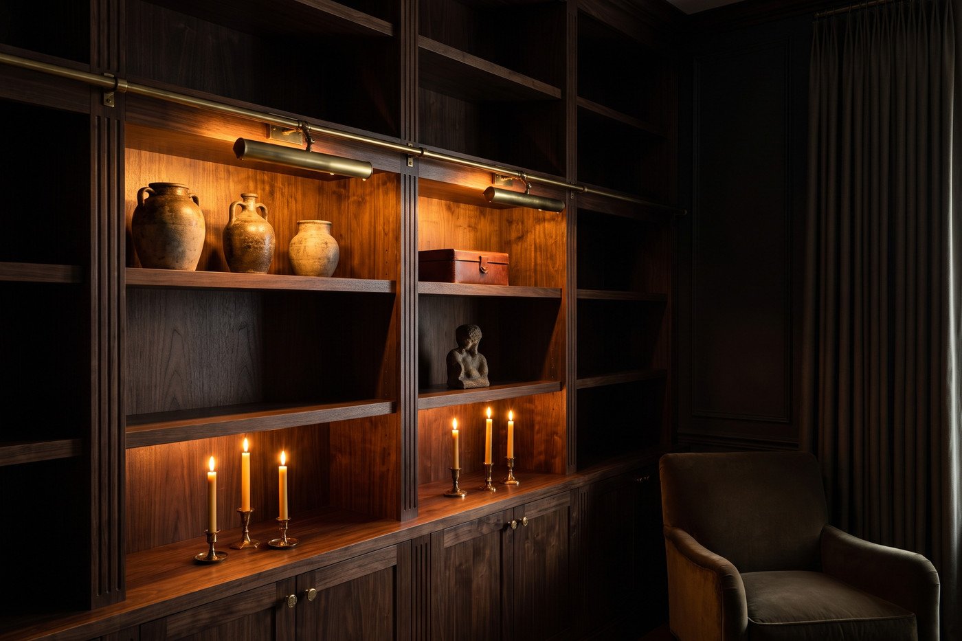 The same bookshelf at night with warm picture lights glowing on two shelves, candles lit below and the rest of the dark room falling away.