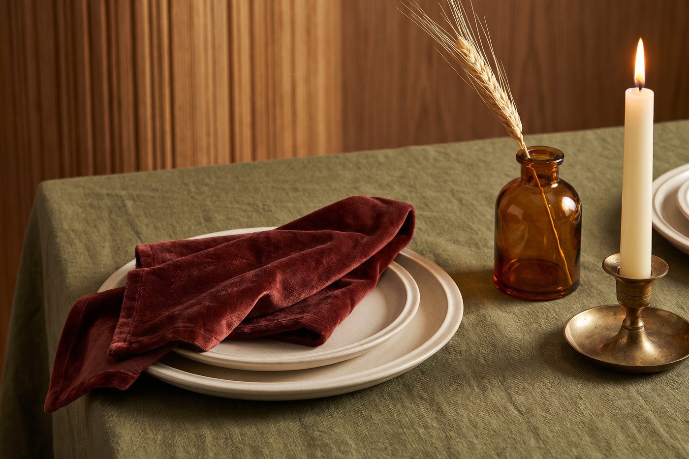 Close view of a fall place setting: cream stoneware on olive linen with an oxblood velvet napkin, an amber bud vase holding dried wheat and a brass candlestick.