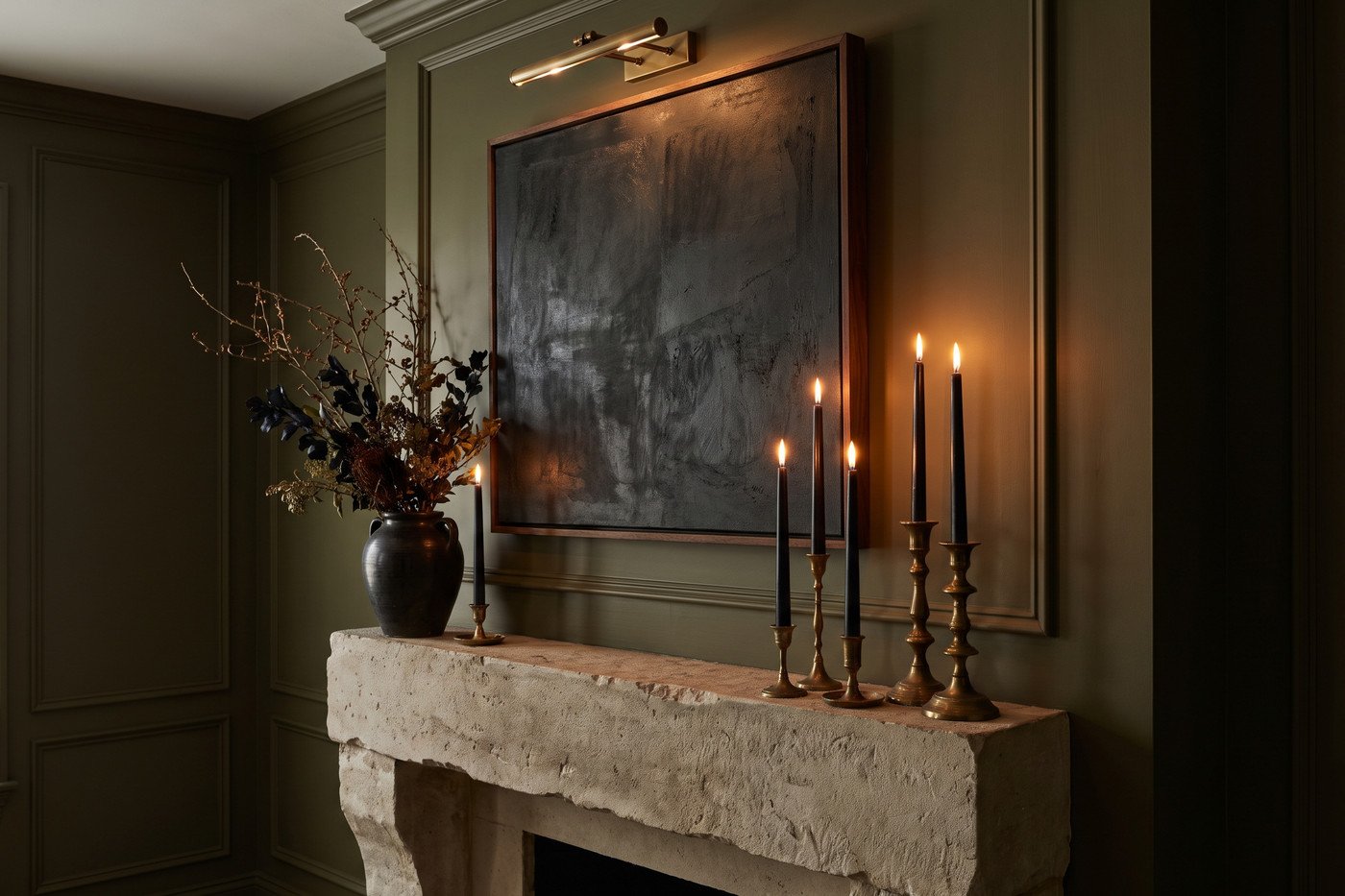 Limestone mantel at night with black taper candles in brass holders at three heights, a black urn of dried dark florals and a moody abstract painting lit by a brass picture light.
