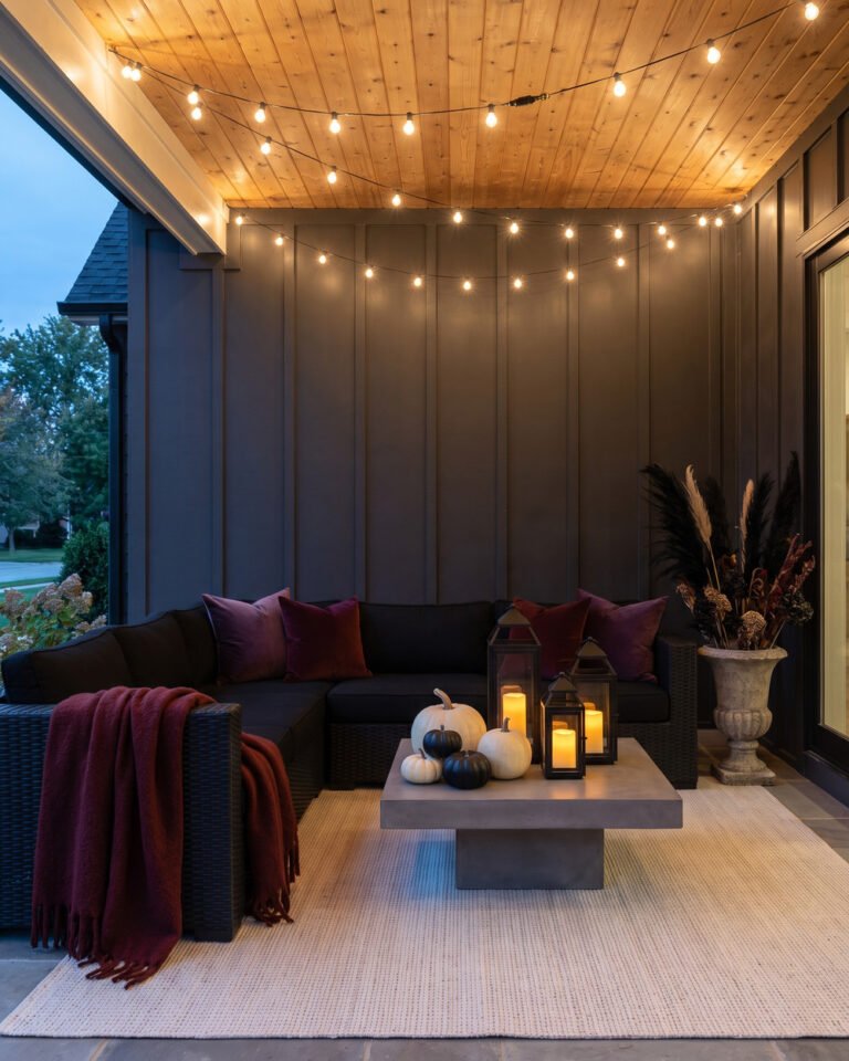 Moody Halloween patio at dusk with a black wicker sectional, plum and oxblood velvet pillows, black lanterns, pale pumpkins and dried dark botanicals against a board-and-batten wall.