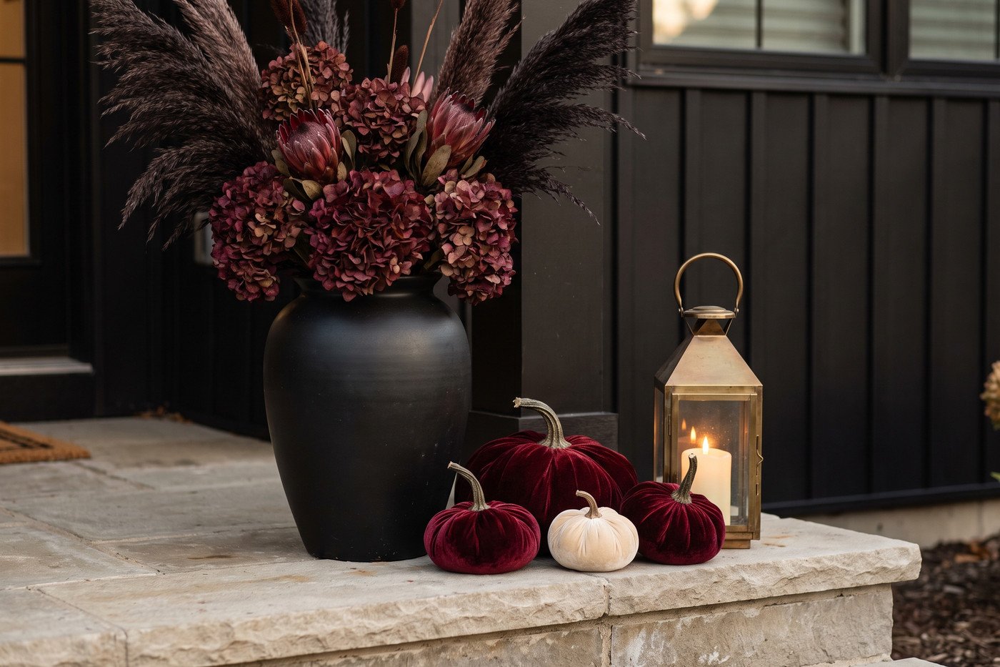 Close view of a black urn planted with oxblood-dyed dried florals and dark pampas beside oxblood velvet pumpkins and a brass lantern on a limestone porch step.