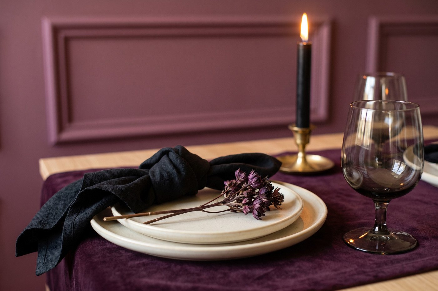Close view of a Halloween place setting: cream stoneware on a plum velvet runner, a black linen napkin, a smoked glass goblet and a sprig of dark dried florals.